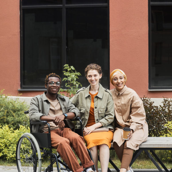 Portrait of smiling young disabled African-American man sitting in wheelchair and posing with multi-ethnic women on bench